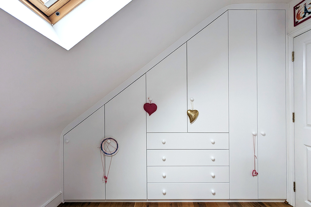 Built-in slanted loft wardrobe in kid's bedroom with 6 doors and drawers in the middle